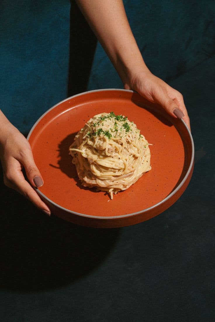 A plate of carbonara held in two hands over a deep blue table